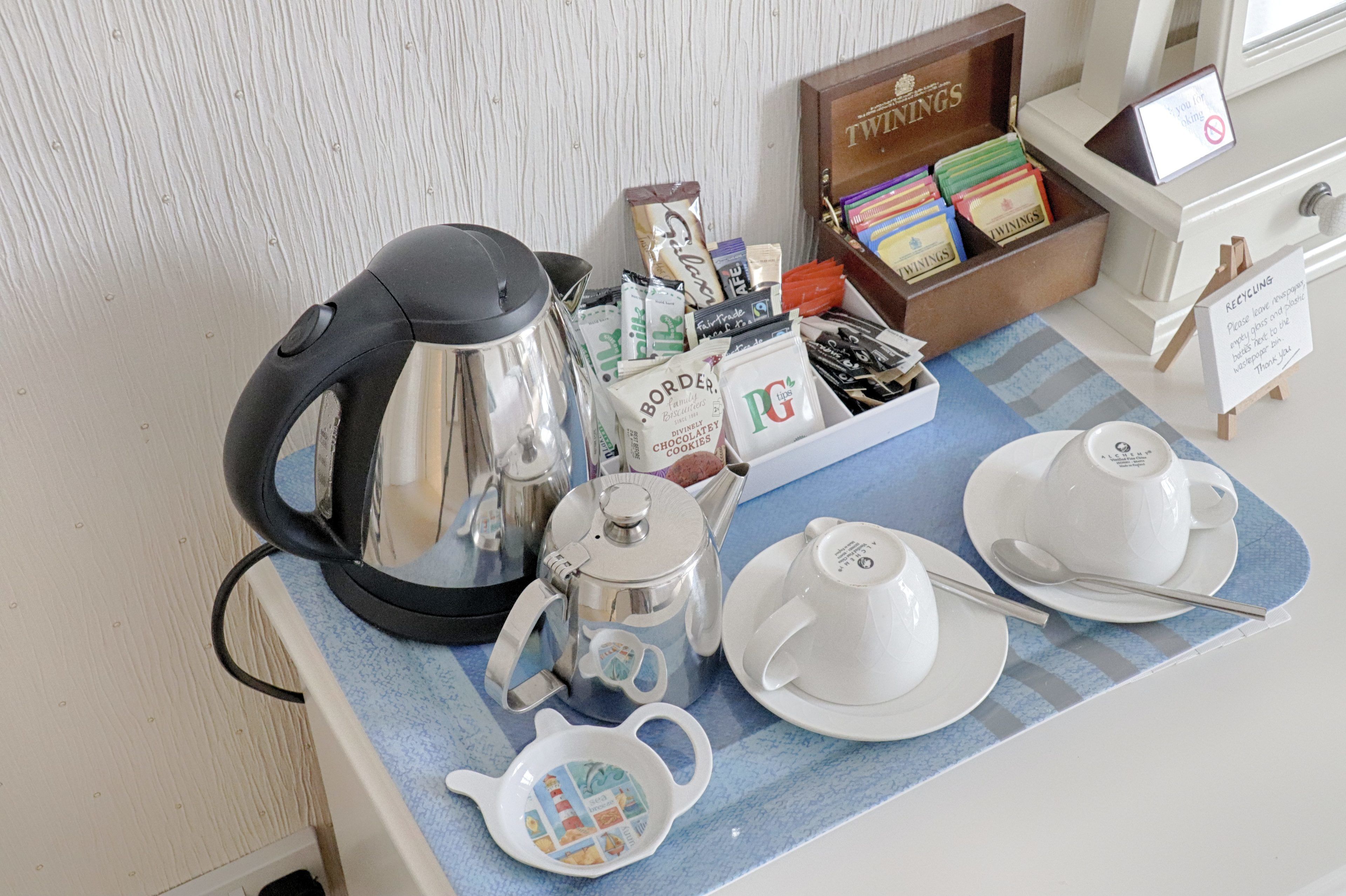 Een ontbijtarrangement met een waterkoker, theepot, kopjes en diverse snacks en toevoegingen op een tafel.