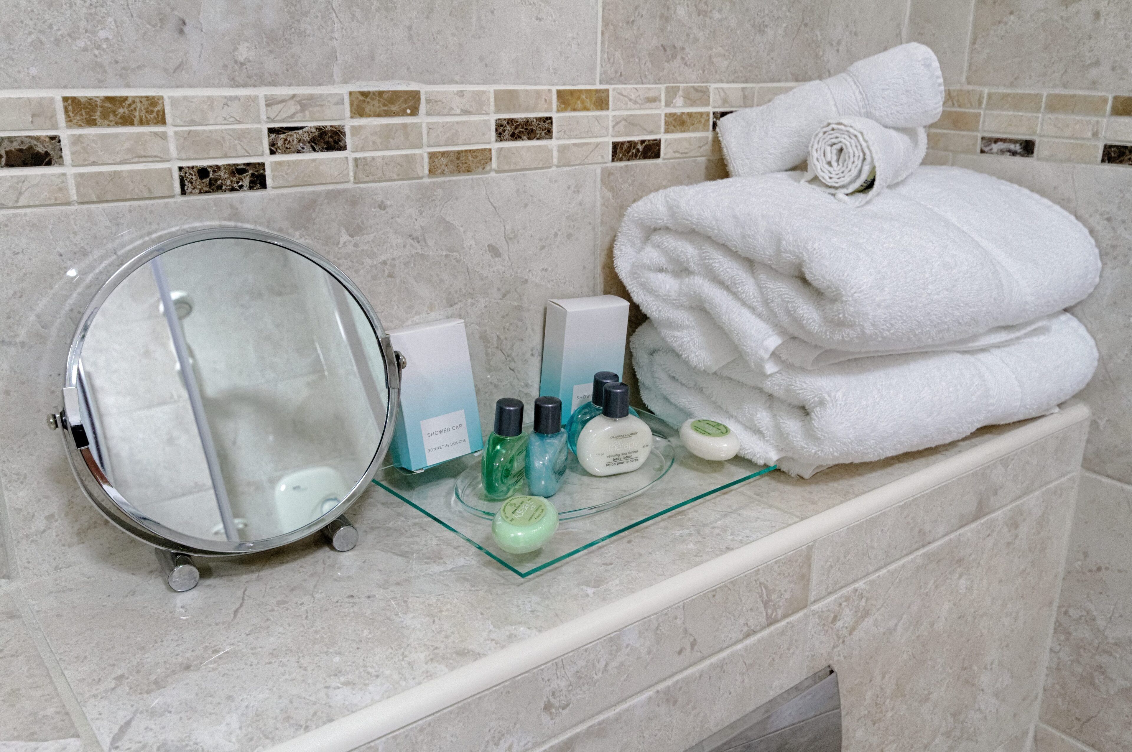 A bathroom vanity with a glass-topped sink, a round mirror, and neatly stacked white towels.