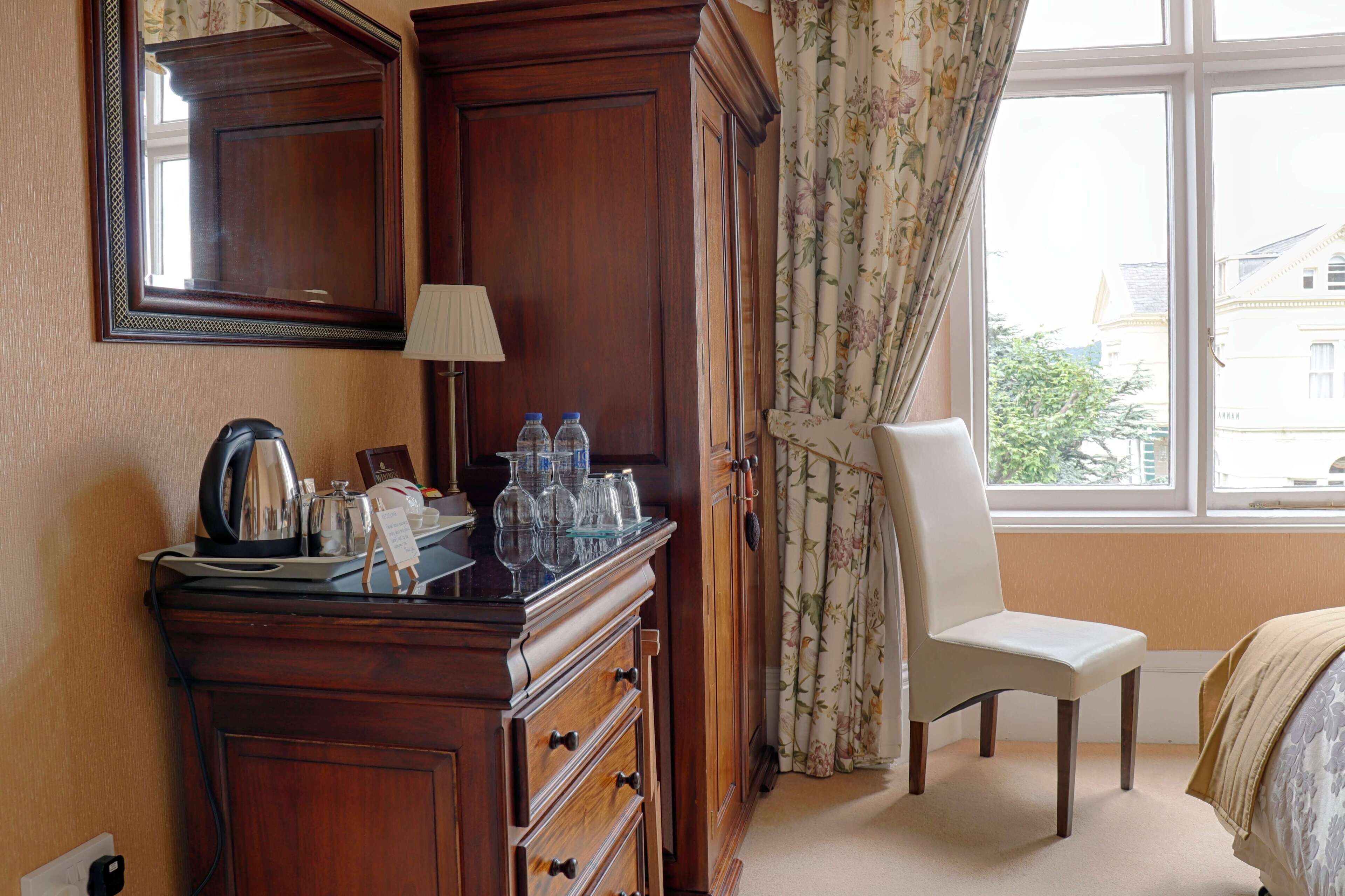 A hotel room with a wooden dresser, a chair, a bed, a window with curtains, and a view of buildings outside.