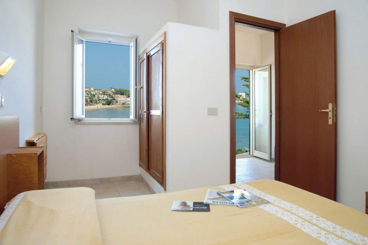 A bedroom with a bed, a window with a view of the sea, and a door leading to an outdoor area.