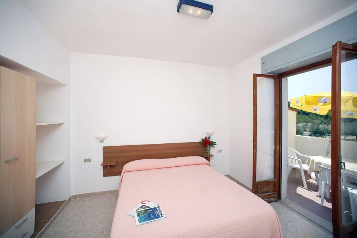 A bedroom with a bed, a wooden wardrobe, a nightstand with a lamp, and a view of a balcony with outdoor furniture.