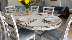 Ein Esstisch ist für sechs Personen gedeckt, mit weißen Tellern, goldfarbenen Gläsern und gewebten Platzsets. Auf dem Tisch steht eine Vase mit gelben Blumen.