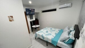 A small, well-lit bedroom with a bed, a small table, and an air conditioning unit on the wall. - Studio D - Queen Room (Chippingham)