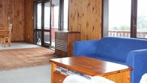 Un interior de madera con un sofá azul, una mesa de madera con libros y un ventanal corredero que da a un espacio exterior.