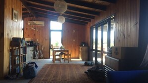 Una habitación rústica con paredes de madera y un ventanal grande. Se observa una mesa de comedor con sillas, una estantería y una silla azul.