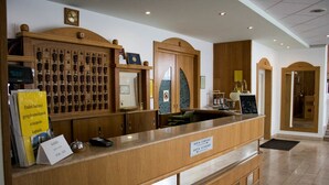 Zona de recepción de un hotel con un mostrador de madera, un reloj digital, una mesa de recepción y un panel en la pared con llaves.