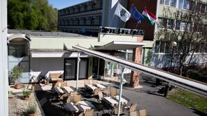 Un área de estar al aire libre con mesas y sillas, un edificio con varias banderas y un cielo despejado.