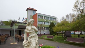 Una estatua en un patio con bancos y un edificio con la leyenda "PLATAN" al fondo.