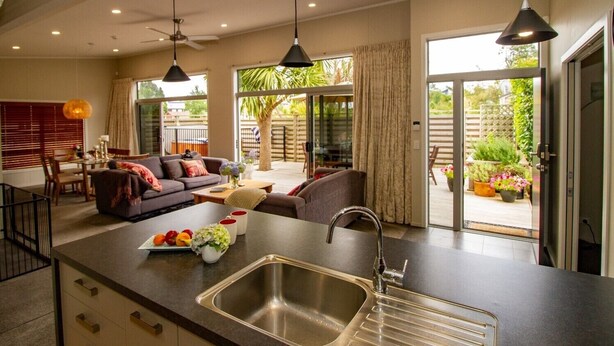 A modern kitchen with a central island, a dining area with a sofa and table, and a patio with a view of greenery. - 1 bedroom Restful retreat w/Spa for couples (Ohakune)