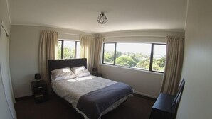 A bedroom with a large bed, a nightstand, a lamp, and a TV. There is a window with curtains and a view of greenery outside. - Skyline Apartments Raglan (Upper Apartment) (Raglan)