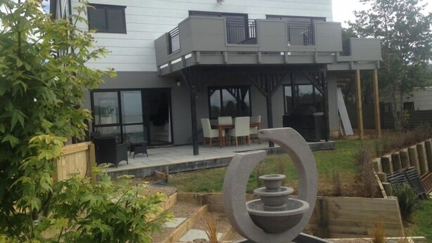 A modern two-story house with a patio area, outdoor seating, and a garden with a decorative stone feature. - Skyline Apartments Raglan (Upper Apartment) (Raglan)