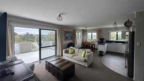 A spacious living area with a sofa, a coffee table, and a dining table. There is a kitchen area with a refrigerator and a sink. Large windows provide a view of the outdoors. - Skyline Apartments Raglan (Upper Apartment) (Raglan)