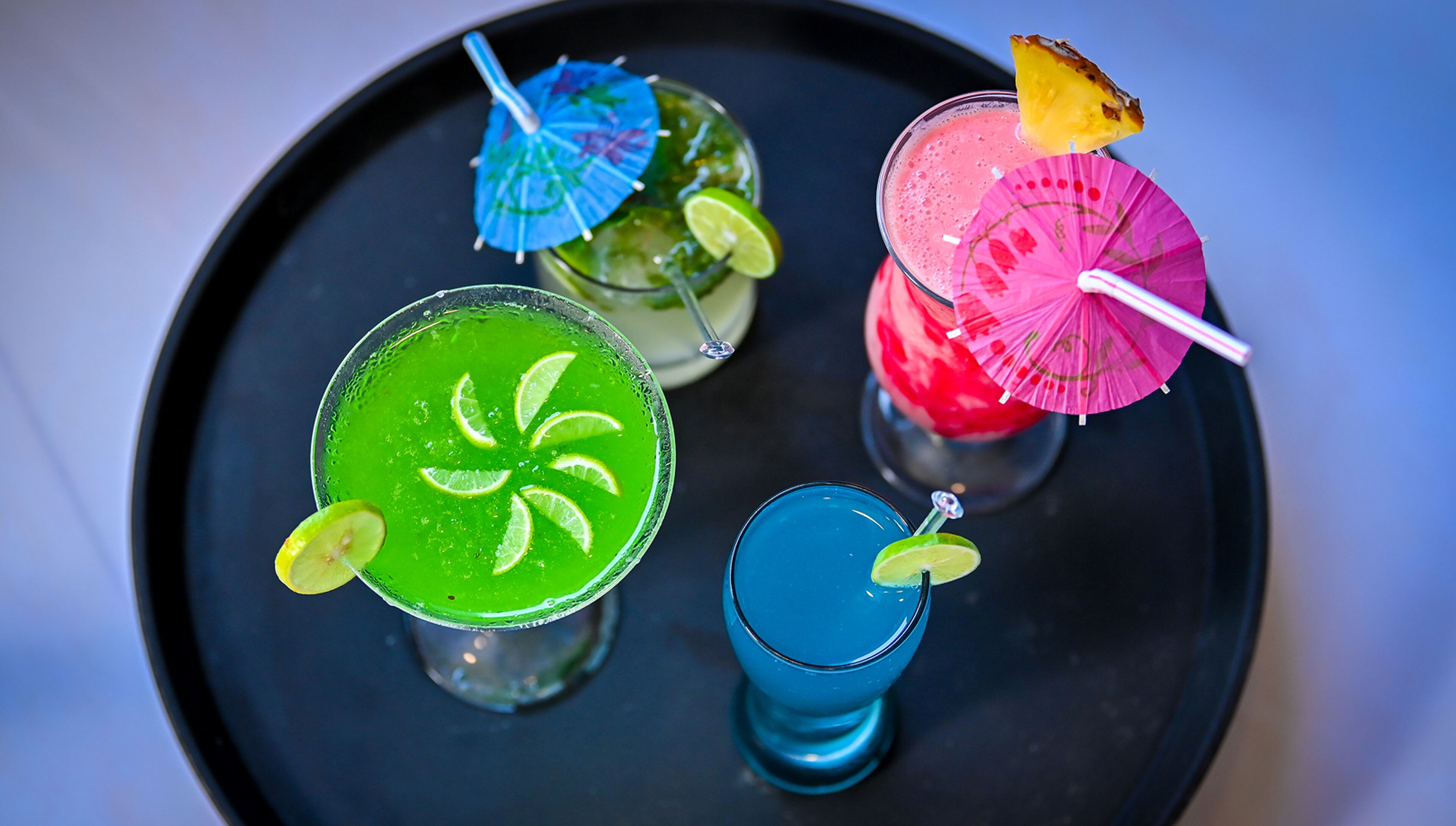 Una bandeja con cuatro bebidas coloridas, cada una adornada con una rodaja de lima y un paraguas decorativo.