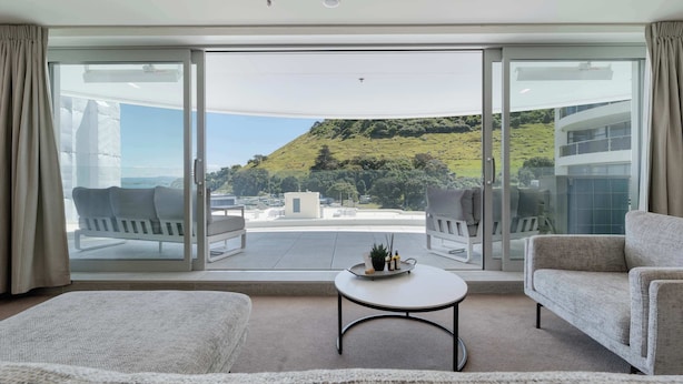 A modern living room with a large sliding glass door leading to a balcony with a scenic view of a hill and a building. - The Oceanside Belle Mt Maunganui (Mt Maunganui)