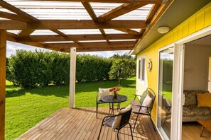 A covered patio with a wooden deck, a table with chairs, and a yellow wall with a sliding glass door leading to an interior space.