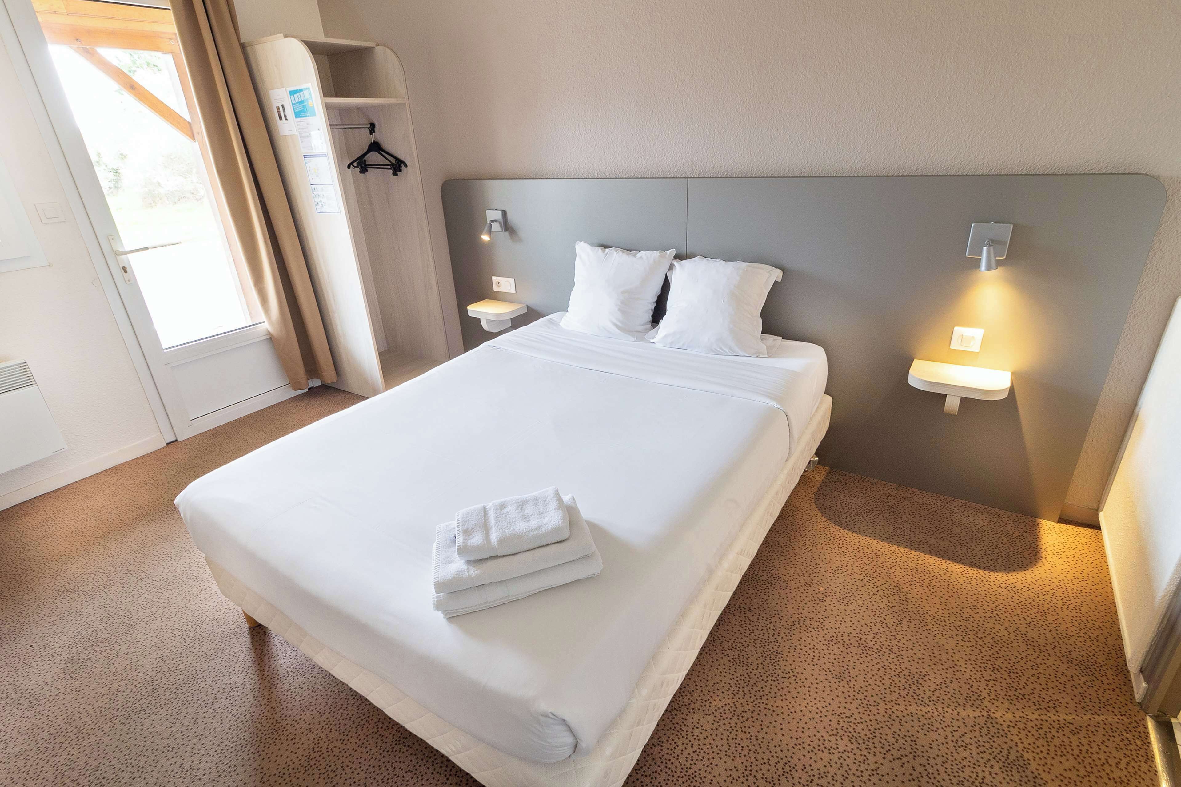 A neatly made bed with white linens and a folded towel, two bedside tables with lamps, a wardrobe, and a door with a view of the outdoors.