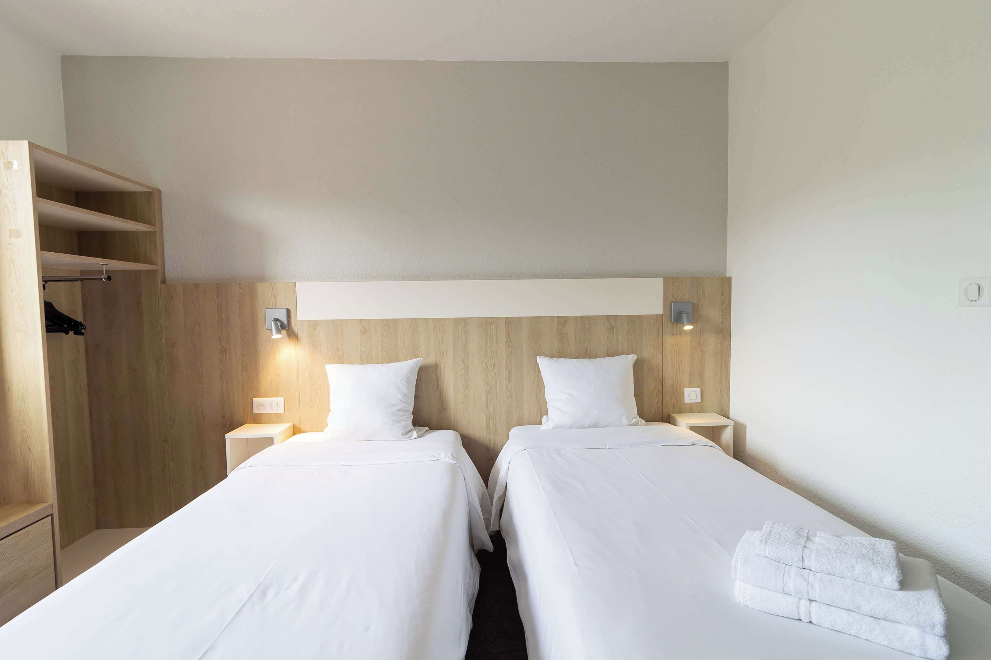 A hotel room with two single beds, a wooden wardrobe, and a small bedside table with a lamp.