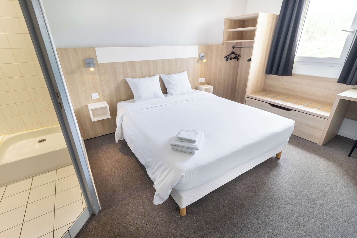 A neatly made bed with white linens and a folded towel, a wooden headboard, a small bedside table, and a window with curtains.