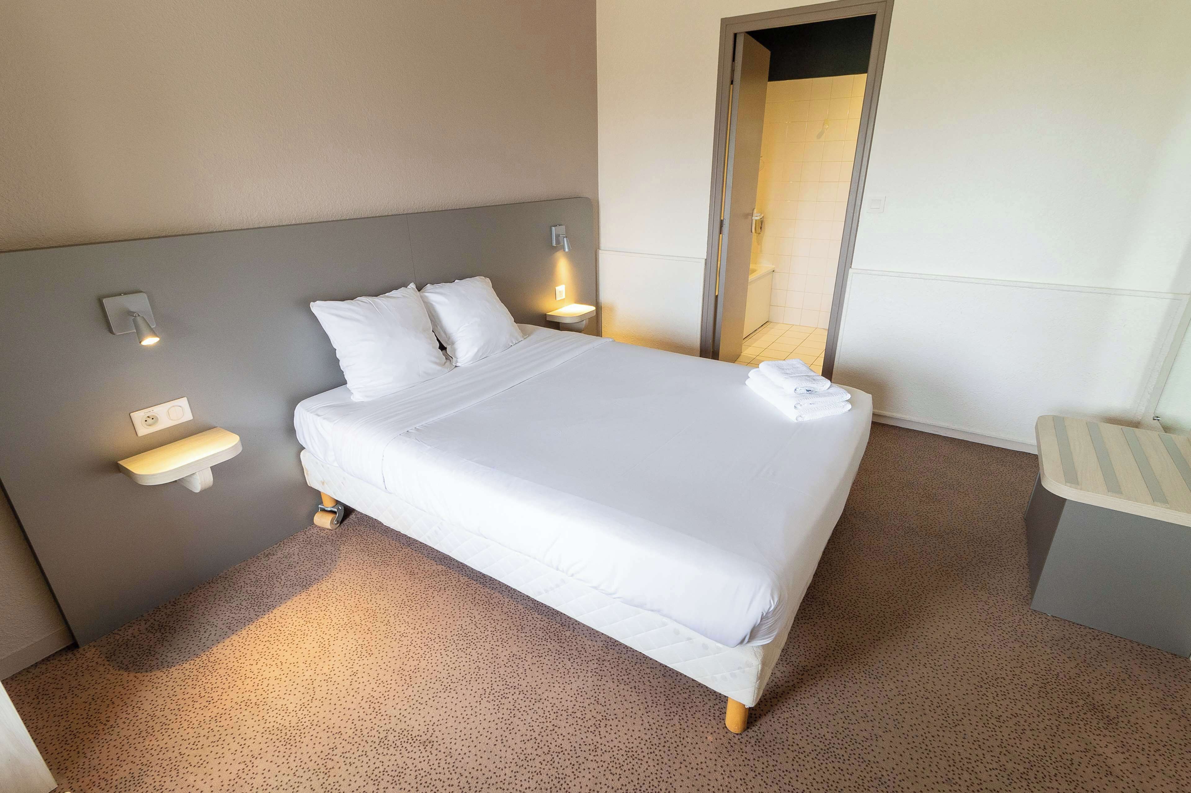 Una cama bien hecha con ropa blanca, una mesita de noche y una lámpara de pared.