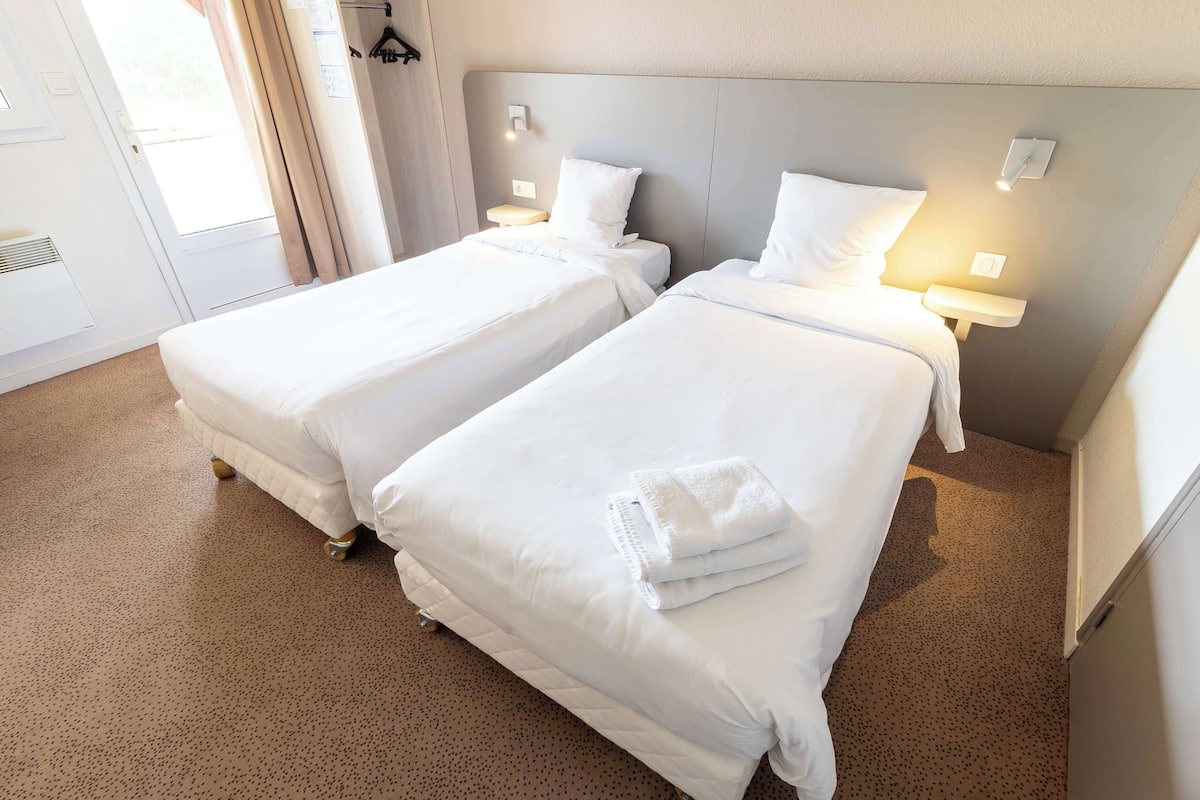 A hotel room with two single beds, each with a bedside table and lamp, and a door with a view.