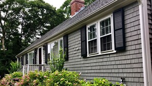A two-story house with a grey siding exterior, black shutters, and a front porch with a small garden. - SPACIOUS & PRIVATE 6 BEDRM HOME CLOSE TO THE OCEAN & BIKE PATH! (Eastham)