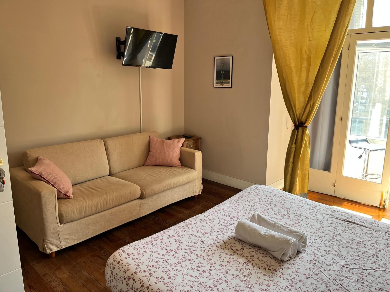 A room with a beige sofa, a flat-screen TV mounted on the wall, a bed with a floral bedspread, and a window with yellow curtains.