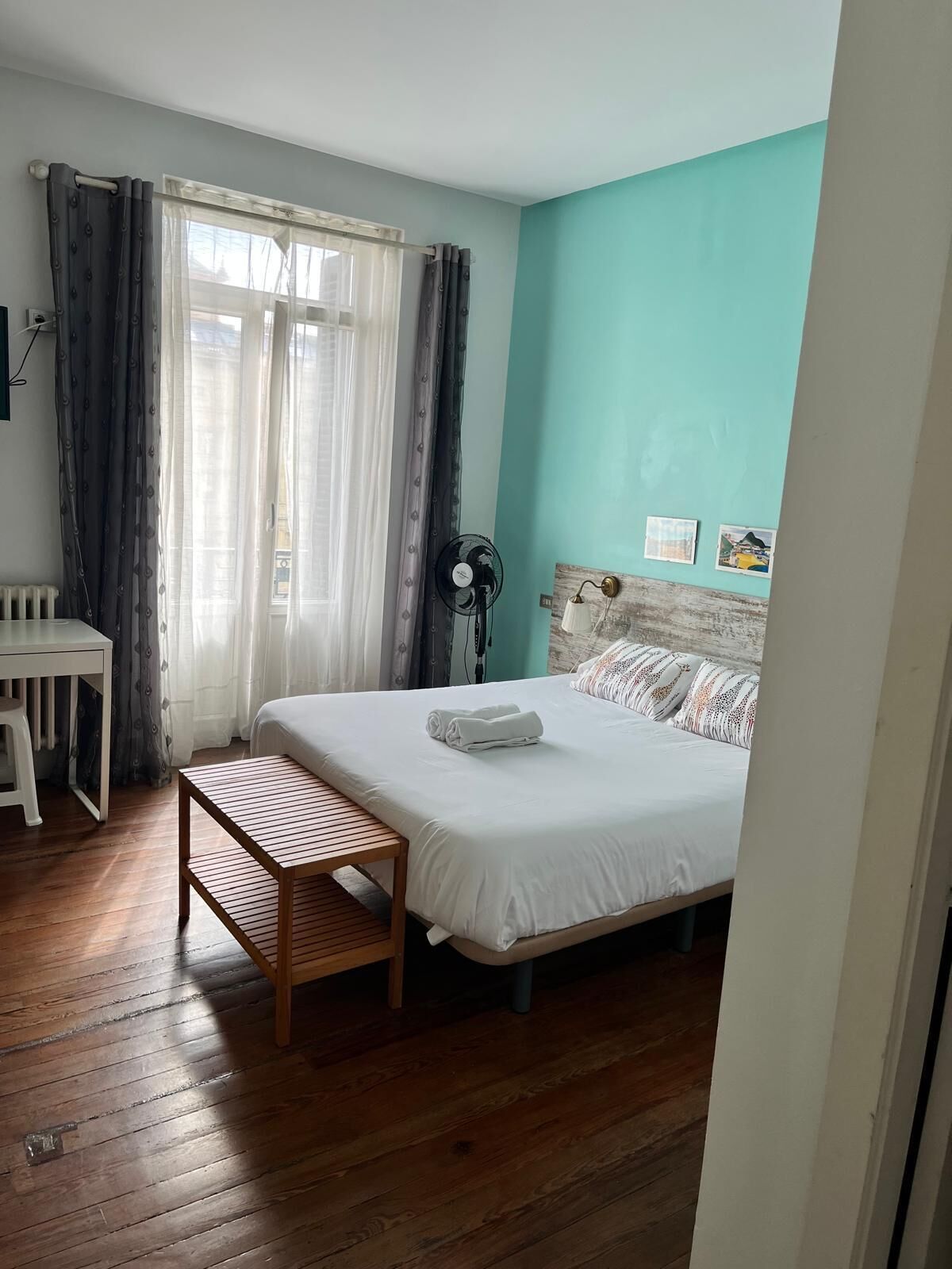 A bedroom with a bed, a bedside table, a chair, a fan, and a window with curtains.