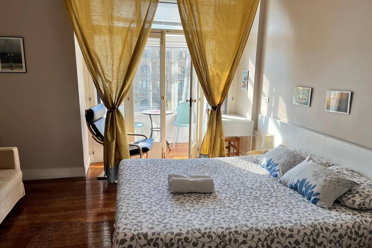 A hotel room with a bed, a chair, a small table, and a view of the outside through a window with curtains.