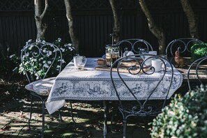 Une table à manger d’extérieur, recouverte d’une nappe à motifs, entourée de chaises en métal, le tout dans un cadre champêtre agrémenté d’arbres et de feuillage.