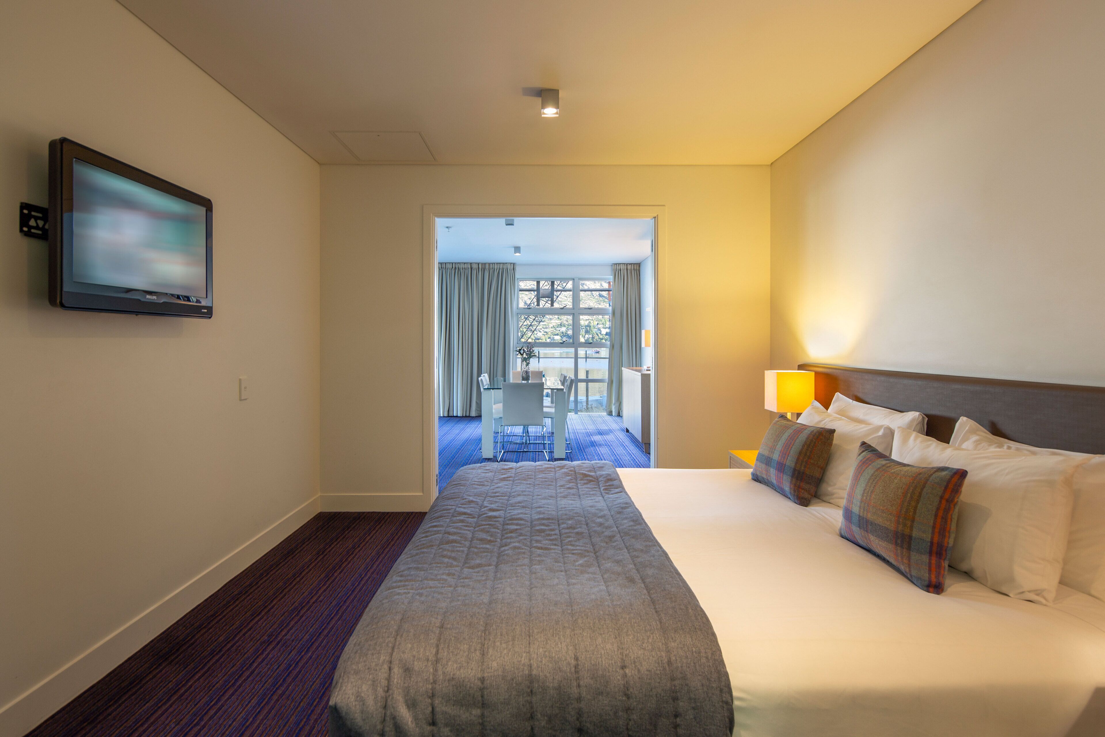 Habitación de hotel con una cama grande, un televisor de pantalla plana en la pared, una mesita de noche con lámpara y vistas al comedor a través de una puerta abierta.