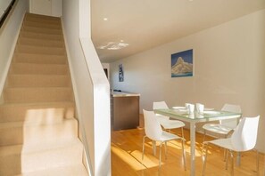 A modern interior with a staircase, a dining area with a glass table, white chairs, and a kitchen in the background.