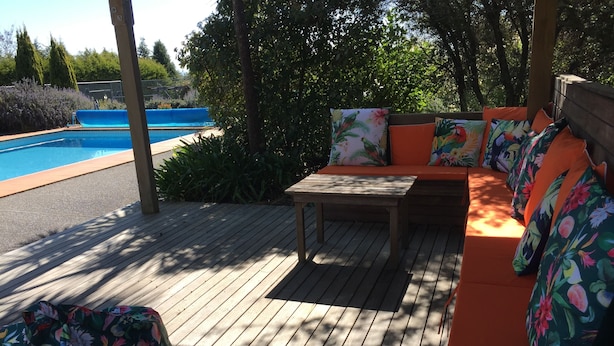 Een houten terras met een zwembad, een tafel en een oranje bank met bloemenkussens.