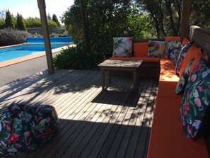 Een houten terras met een zwembad, een tafel en een oranje bank met bloemenkussens.