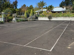 An outdoor tennis court with a net, surrounded by trees and a building in the background. - Ambury House - private garden resort with views (Havelock North)