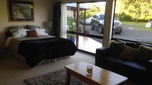 Een slaapkamer met een bed, een houten bijzettafeltje met een glas, een donkergrijze bank en een groot raam met uitzicht op geparkeerde auto's.