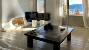 A room with a wooden floor, a blue sofa with cushions, a stone fireplace, a wooden coffee table, and a view of the sea through a window. - Villa Savannah (Mykonos)