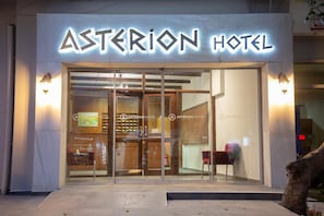 Exterior del hotel Asterion, con entrada de vidrio y señalización iluminada.