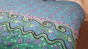Un lit recouvert d’une couvre-lit bleue et verte ornée de motifs représentant des étoiles de mer, des poissons et des coquillages.