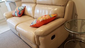 Un sofá de cuero color beige con dos almohadones de colores y una mesa de centro de vidrio.