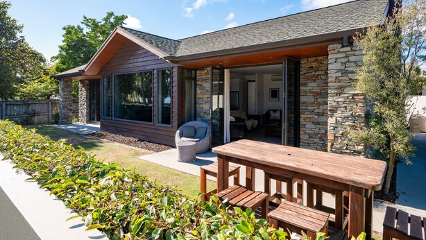 Une maison moderne avec une façade en pierre, de grandes fenêtres et un patio extérieur doté d’une table et de chaises en bois.