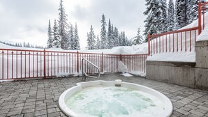 A hot tub in a snowy outdoor area with a red fence and snow-covered trees in the background.