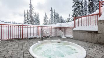 A hot tub in a snowy outdoor area with a red fence and snow-covered trees in the background.