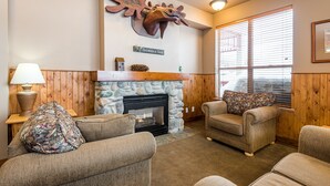 A cozy living room with a stone fireplace, wooden walls, and a mounted deer head sculpture. There are two armchairs and a lamp on a side table.