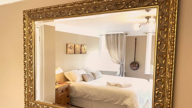 A bedroom with a large ornate mirror, a bed with white linens, a nightstand with a lamp, and a wall-mounted artwork.