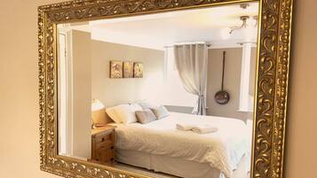 Une chambre à coucher avec un grand miroir décoratif, un lit recouvert de draps blancs, une table de chevet avec une lampe et une œuvre d’art accrochée au mur.