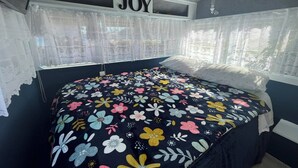 Quarto com colcha florida, um letreiro 'JOY' sobre a cama, cortinas leves e vista para o verde lá fora.