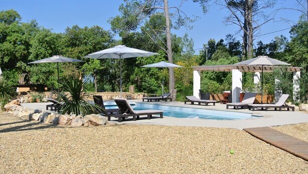 Un espace piscine agrémenté de chaises longues et de parasols, entouré d’arbres et surmonté d’une pergola.