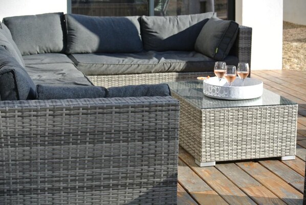 Un área de estar exterior moderna con un sofá trenzado gris y una mesa de centro de vidrio con bebidas.