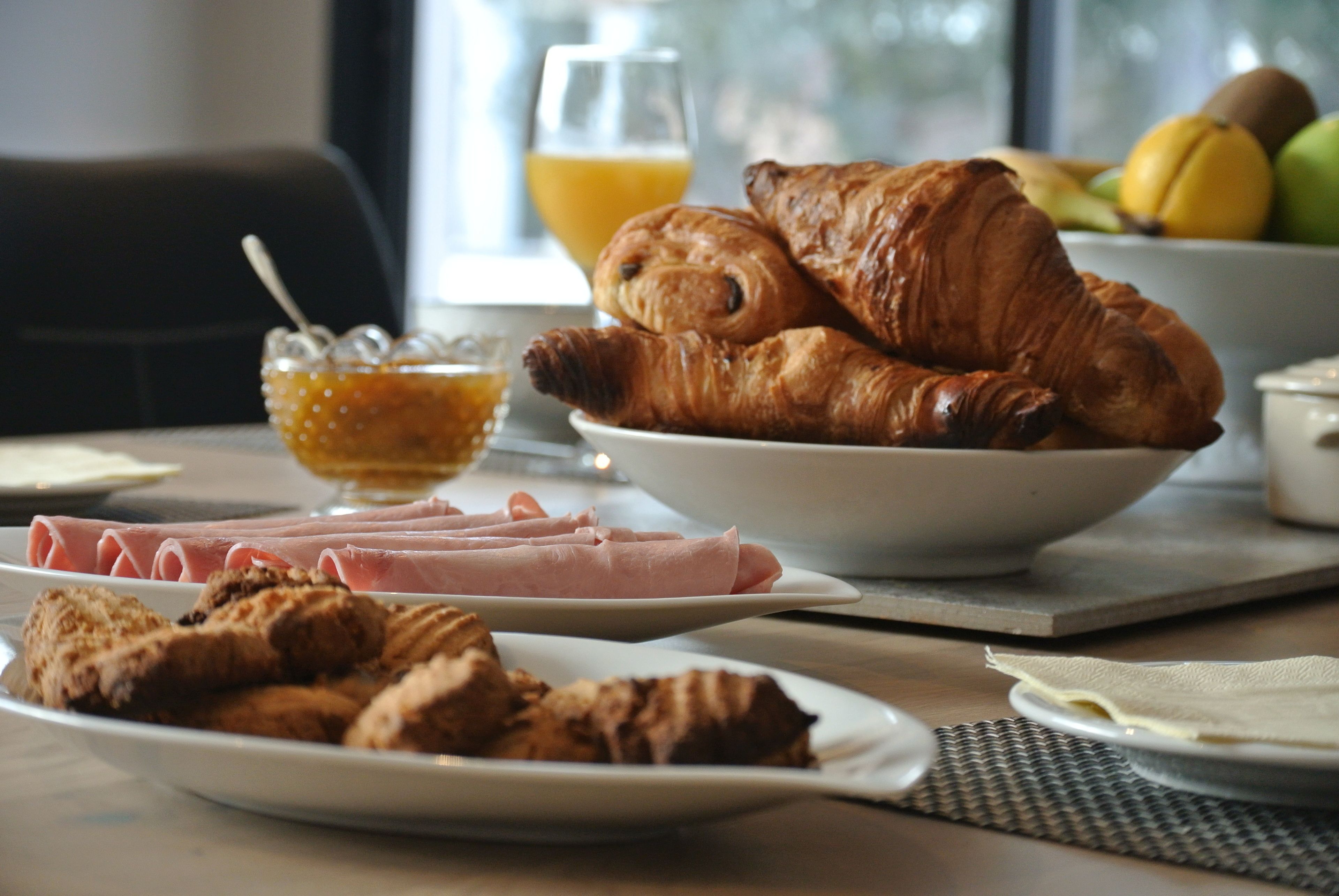 Una mesa de desayuno con croissants, jamón y un bol de fruta.