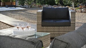 Un espace de détente extérieur aménagé avec des meubles en osier, comprenant un banc rembourré et une table en verre sur laquelle sont posés des verres à vin.